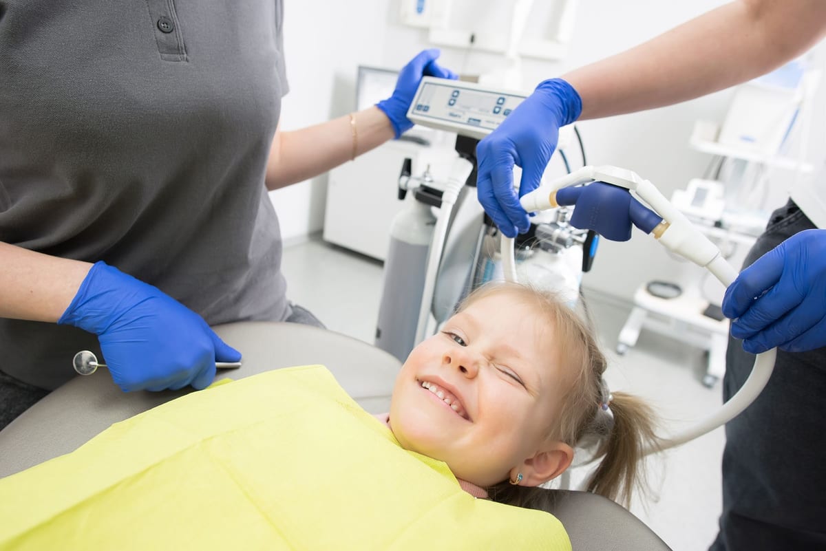 A hygiene team cleaning a young patient's teeth in a bright treatment room